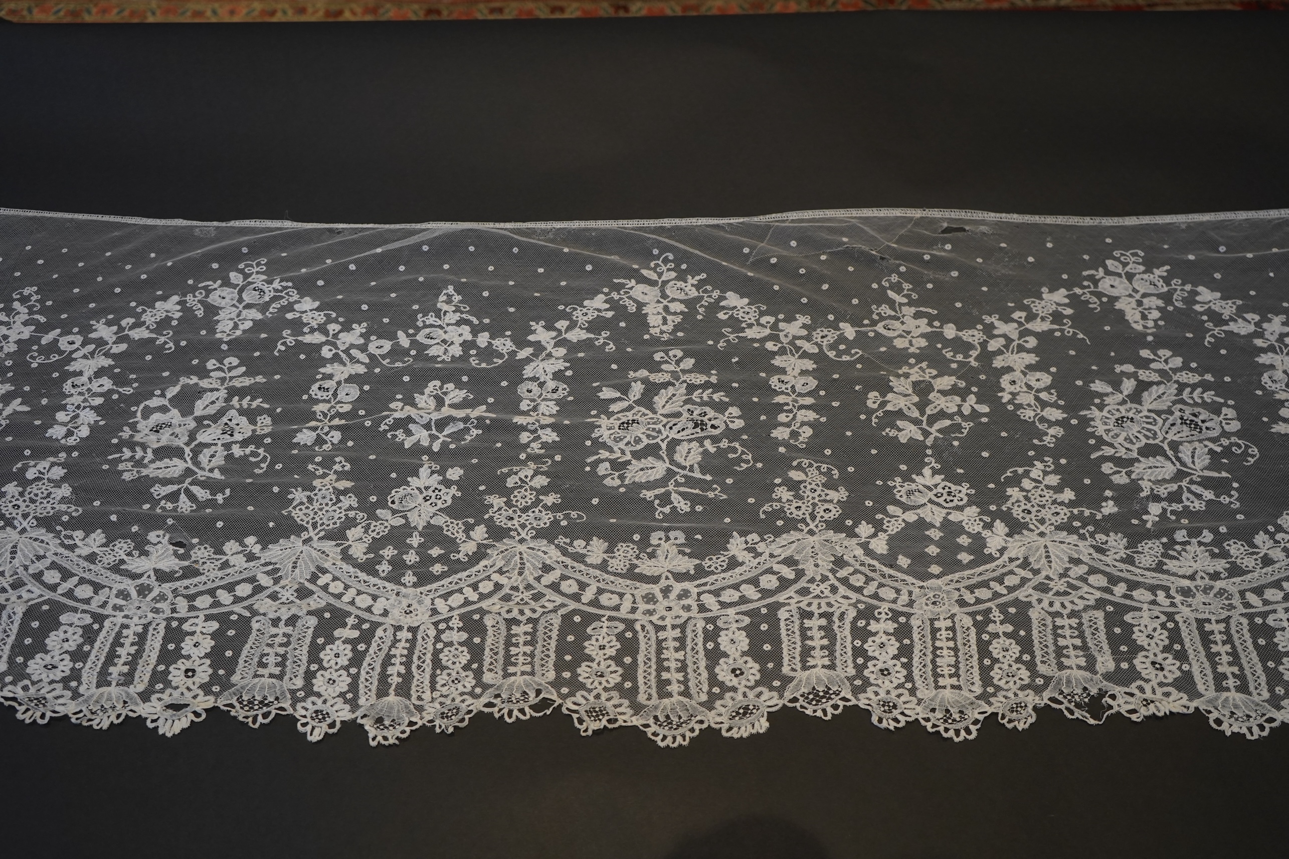 A deep elaborately designed flounce of mid 19th century Brussels bobbin appliquéd lace on net, with finished lace ends, worked with an intricate floral border below large floral cartouches framed in similar motifs and al